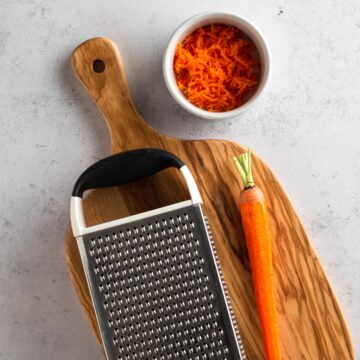 Box grater and peeled carrot on a wooden cutting board.
