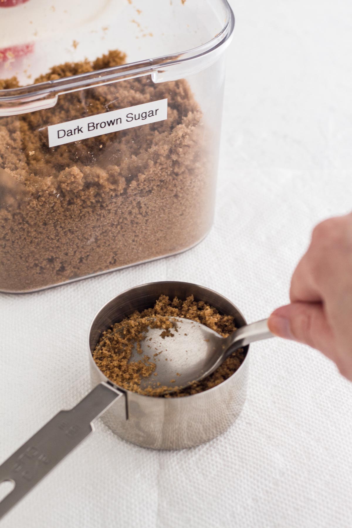 A hand pressing the back of a spoon into a measuring cup to pack down brown sugar.