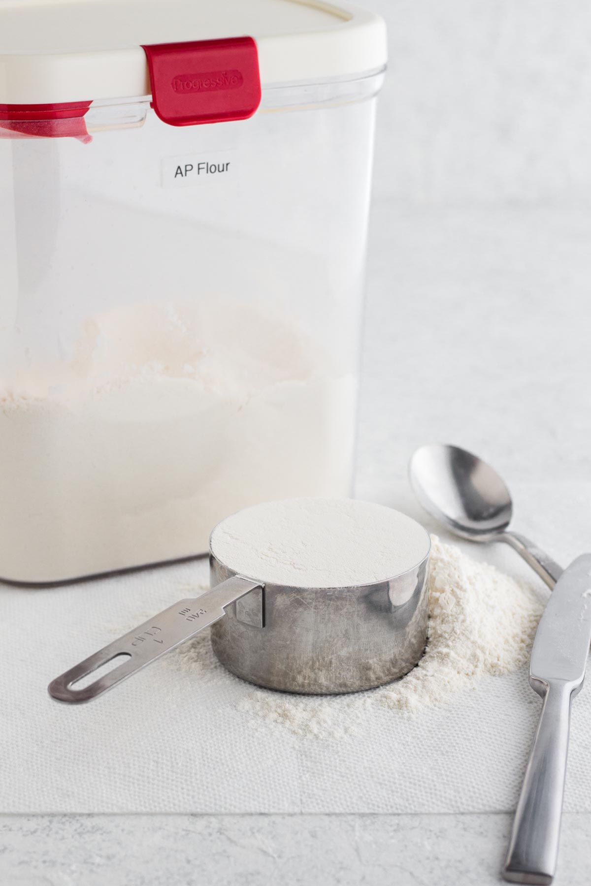 Flour in a measuring cup with a spoon, butter knife, and flour canister on a white surface.