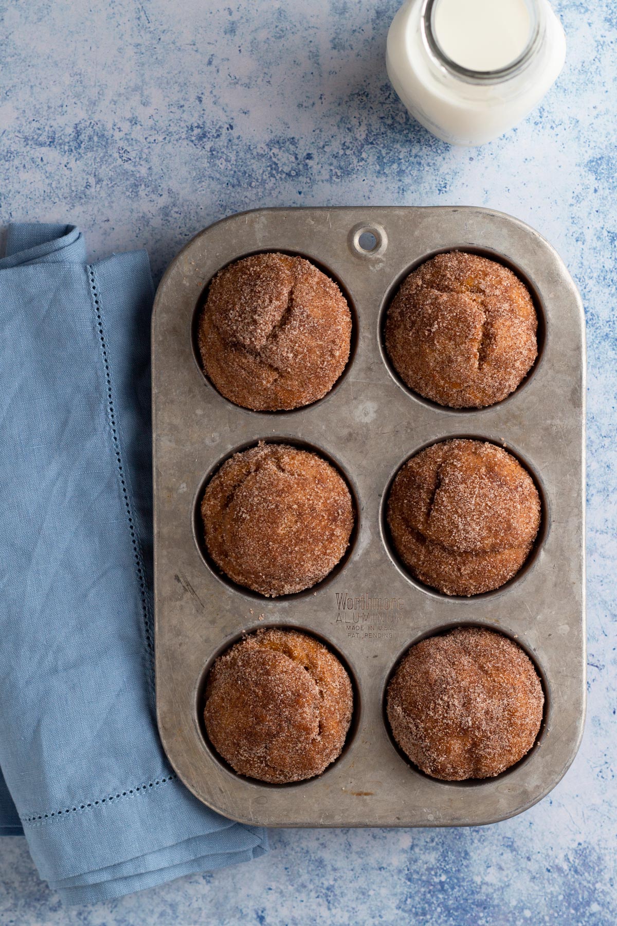 Cinnamon sugar-coated donut muffins in a muffin pan on a blue surface with a napkin and glass of milk.