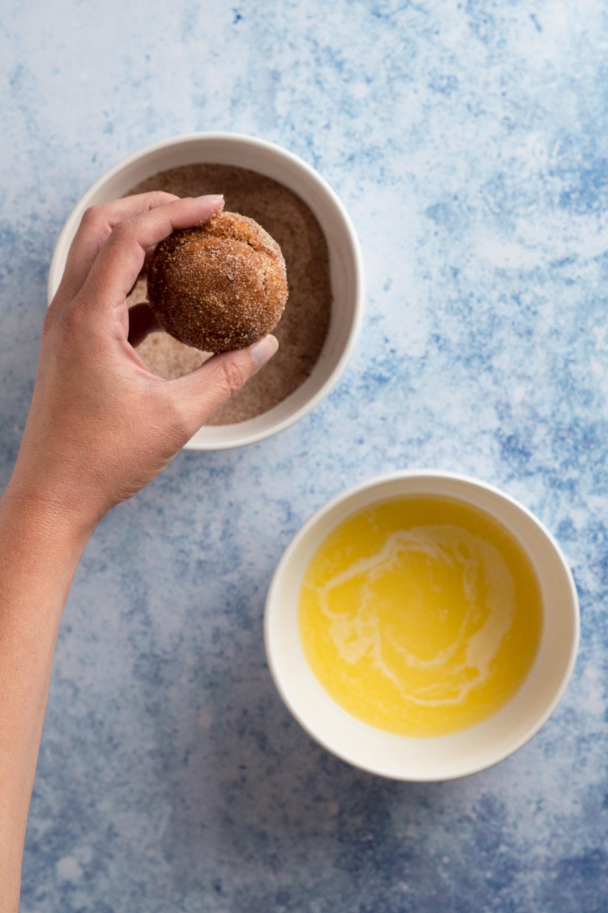 Hand holding donut muffin coated in cinnamon sugar with bowls of melted butter and cinnamon sugar.