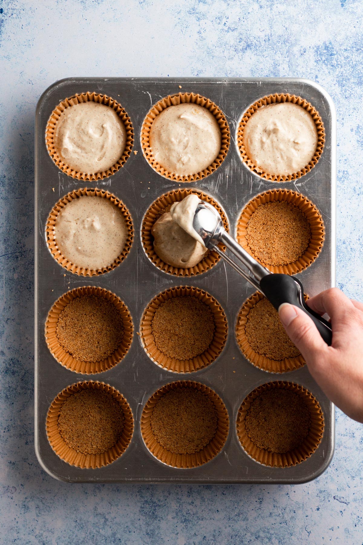 Hand scooping cheesecake mixture onto baked crusts in muffin pan.
