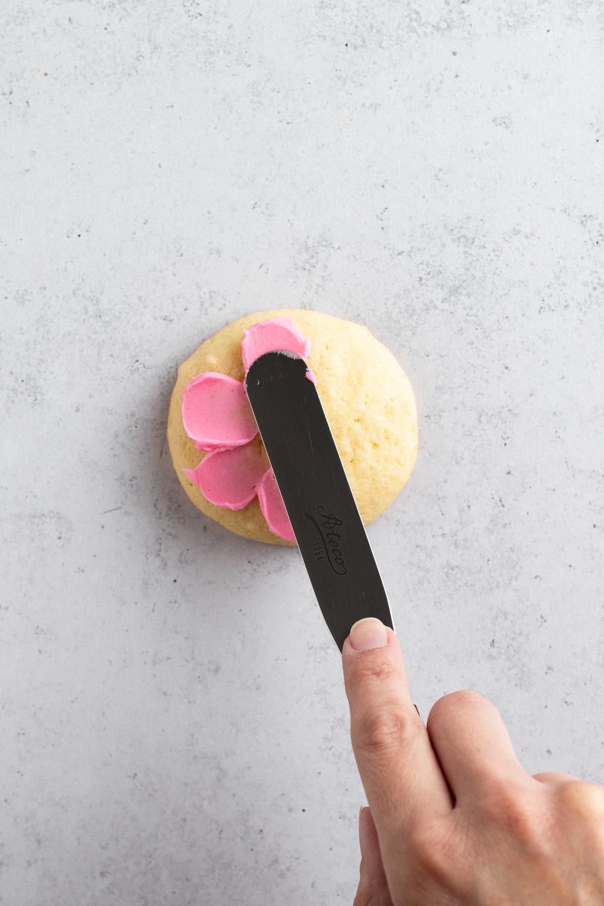 Hand using flat spatula to "paint" pink buttercream flower petals onto a sugar cookie.