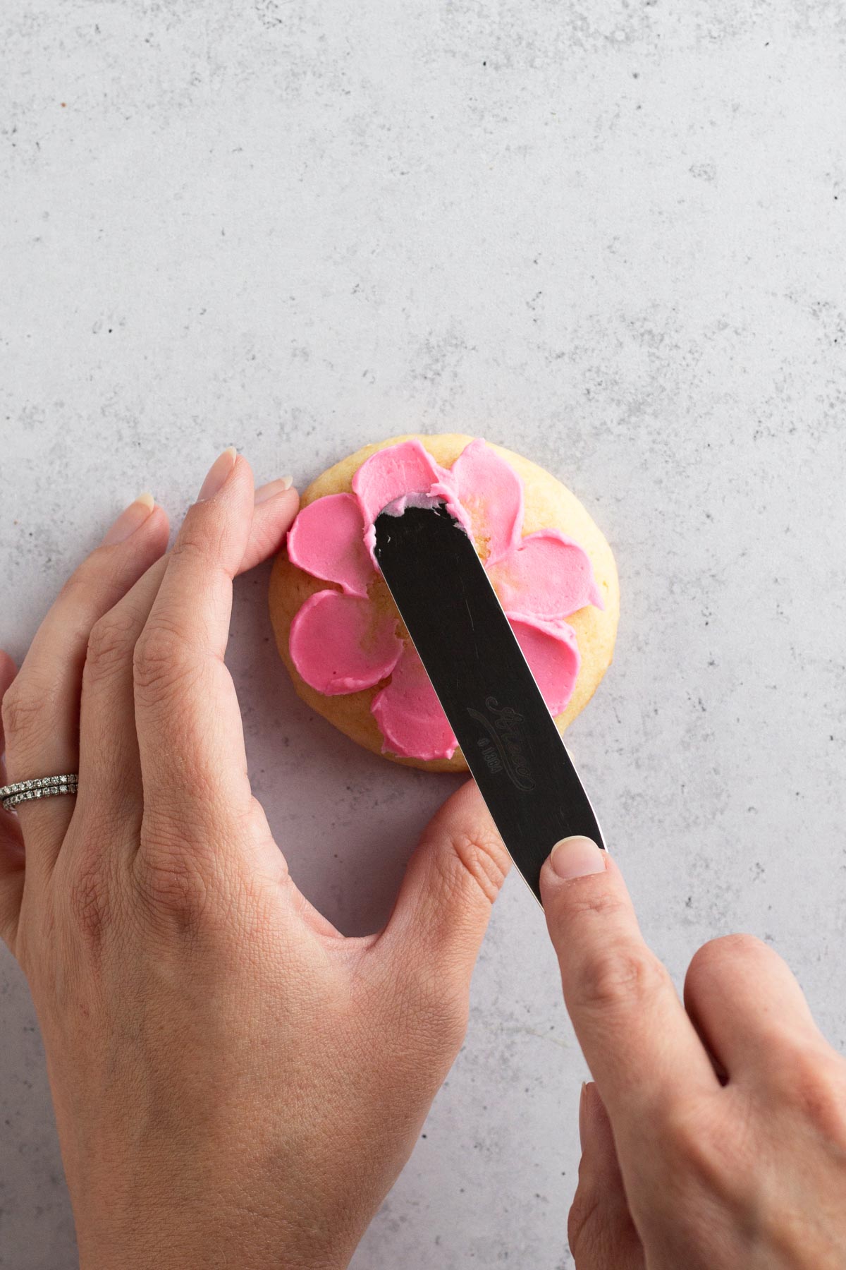 Hands using flat spatula to "paint" pink buttercream flower petals onto a sugar cookie.