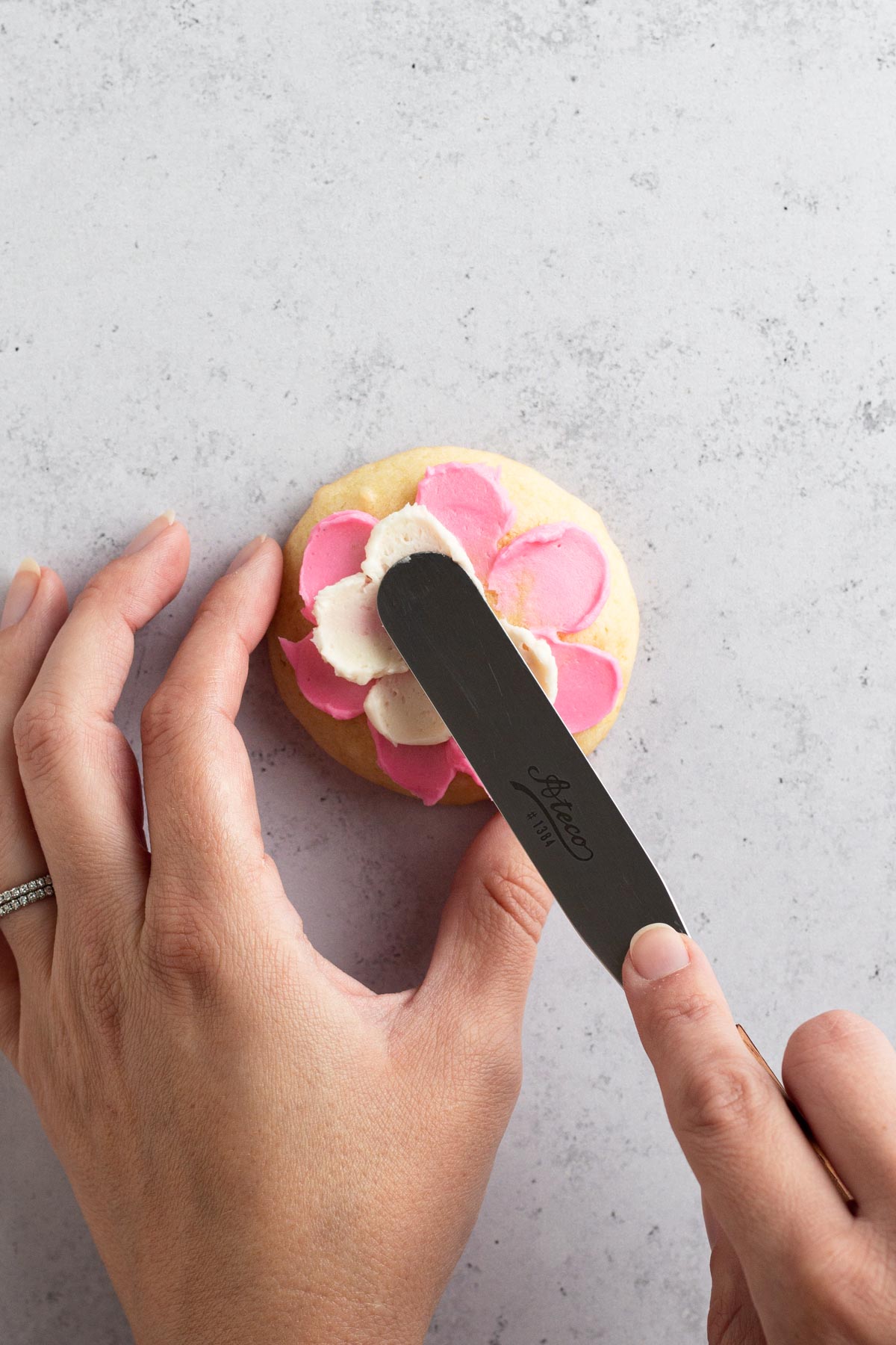 Hands using flat spatula to "paint" white buttercream flower petals onto a sugar cookie.