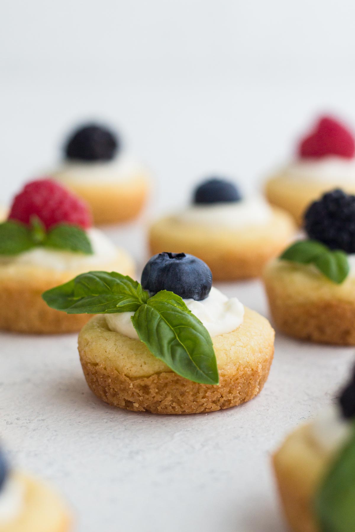 Mini fruit tarts topped with blueberry and a basil sprig on a white surface.