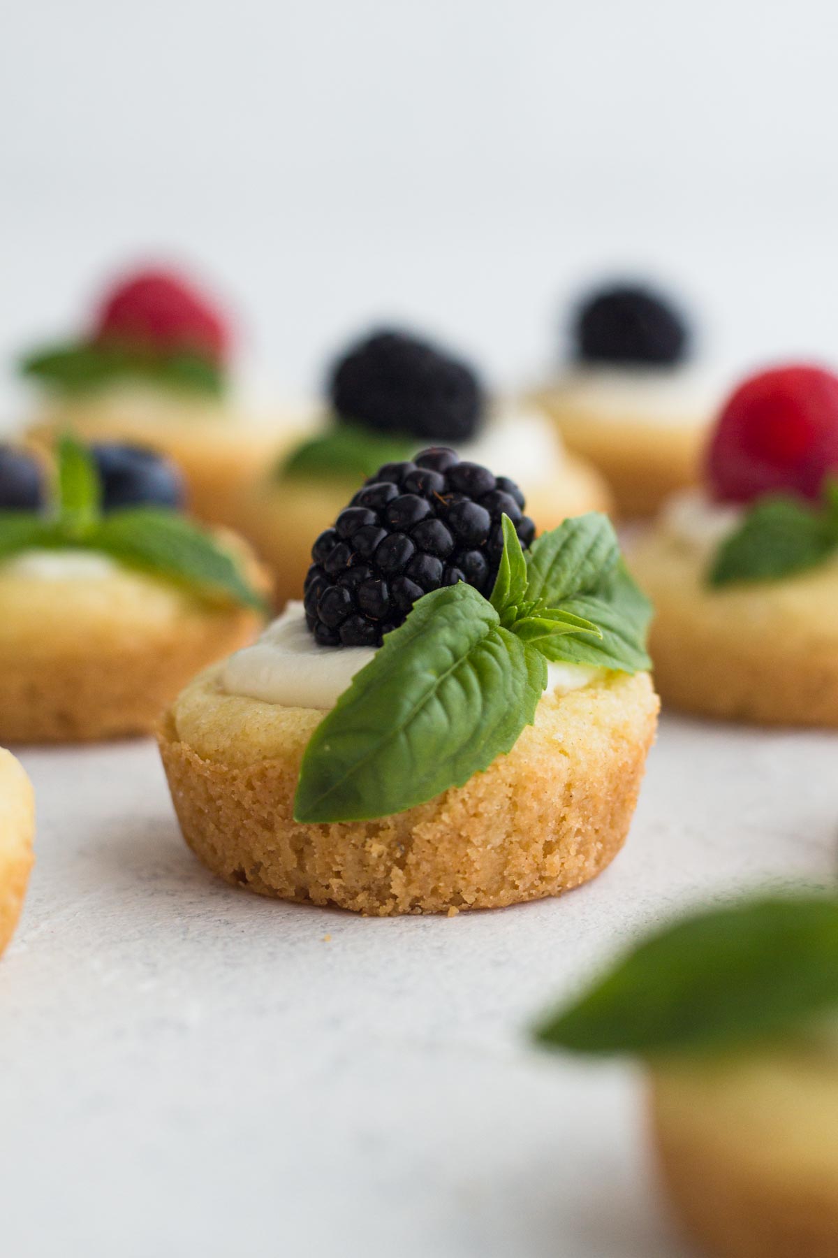 Mini fruit tarts topped with blackberry and a basil sprig on a white surface.