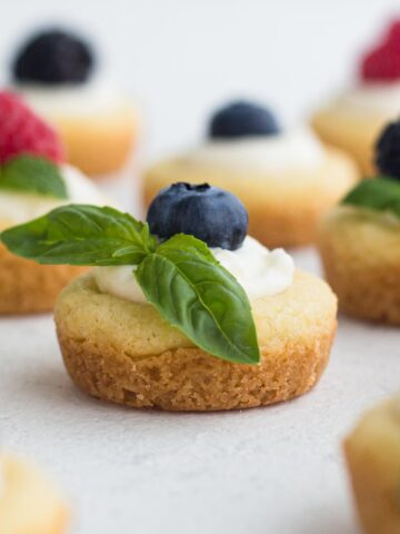 Mini fruit tarts topped with blueberry and a basil sprig on a white surface.