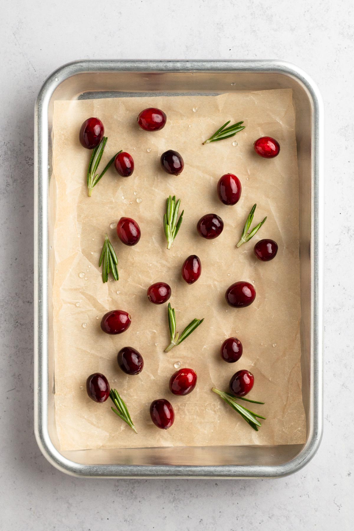 Simple syrup-soaked cranberries and rosemary sprigs on a baking sheet lined with parchment paper.
