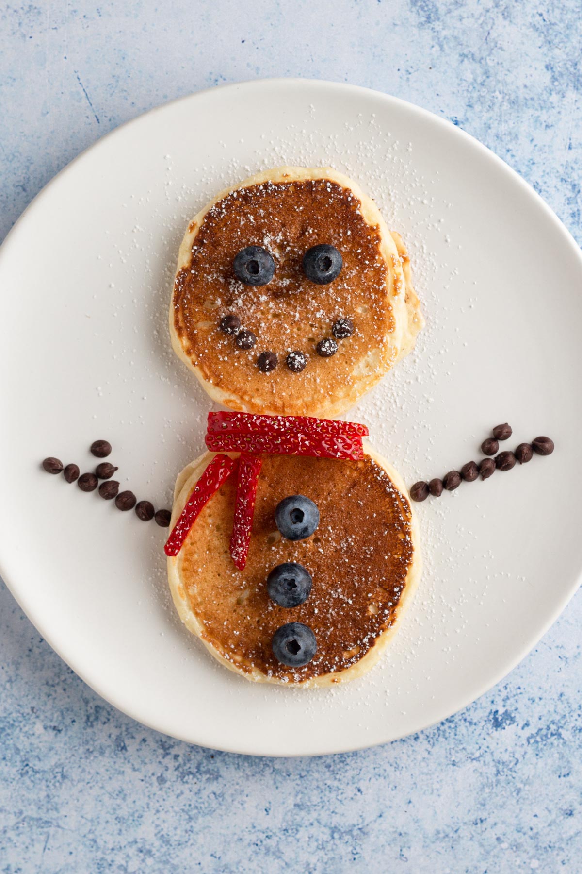 Pancakes on a white plate decorated like a snowman with berries and mini chocolate chips.