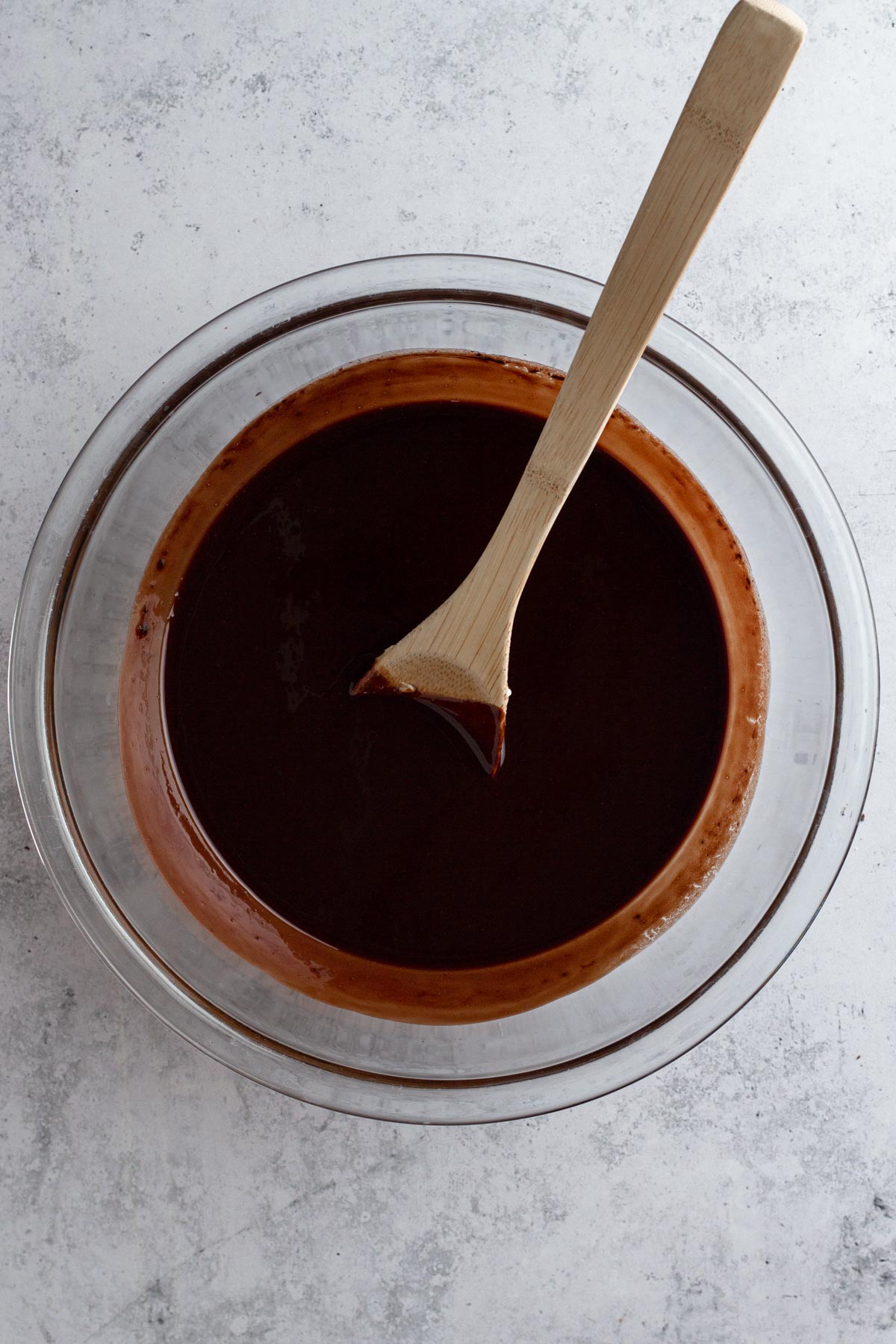 Melted chocolate and butter blended together in a glass bowl with a wooden spoon.