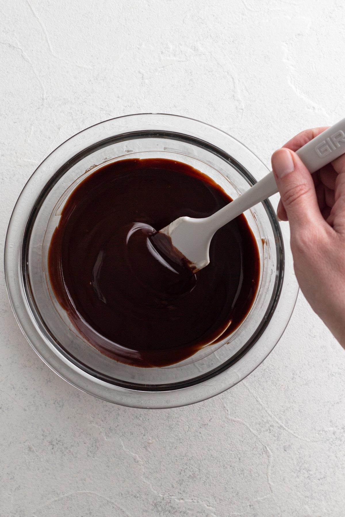 Hand stirring chocolate ganache in a glass mixing bowl using a rubber spatula.