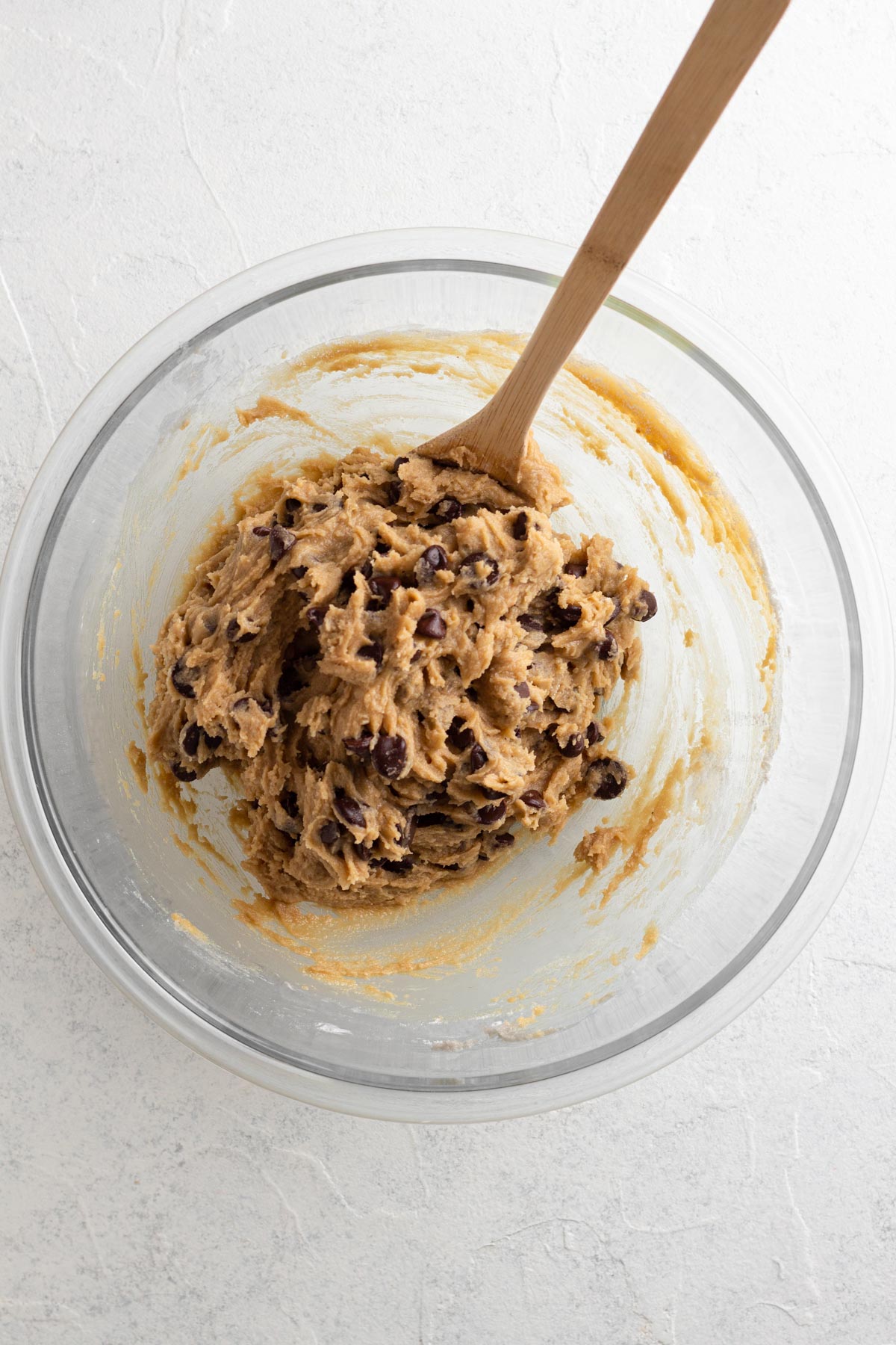 Cookie dough with chocolate chips in a glass mixing bowl with a wooden spoon.