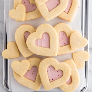 Heart shaped cookies with pink buttercream frosting stacked on a baking sheet.