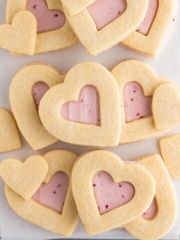 Heart shaped cookies with pink buttercream frosting stacked on a baking sheet.