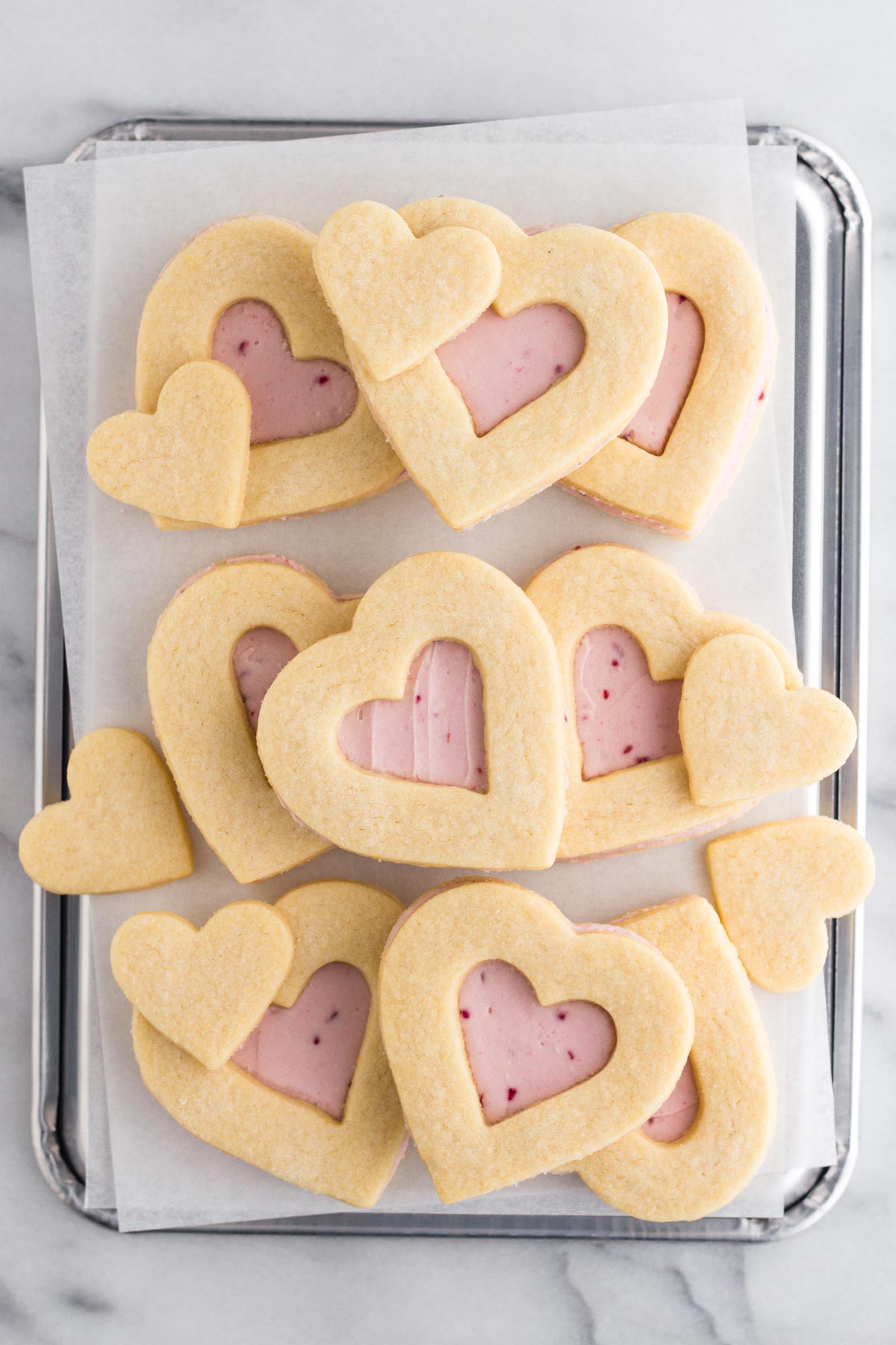 Heart shaped cookies with raspberry buttercream frosting stacked on a baking sheet.
