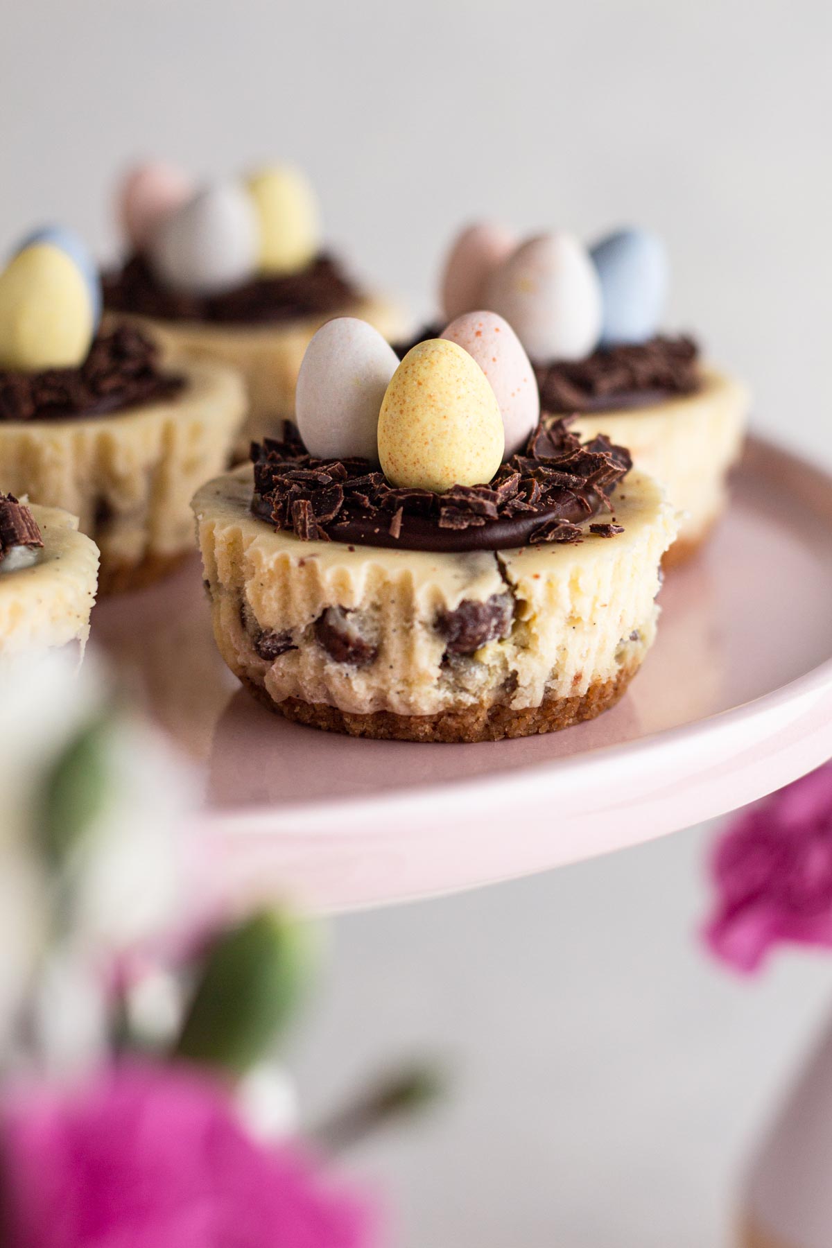 Mini cheesecakes topped with chocolate "nests" and Cadbury mini eggs on a pink cake stand with pink and white flowers.