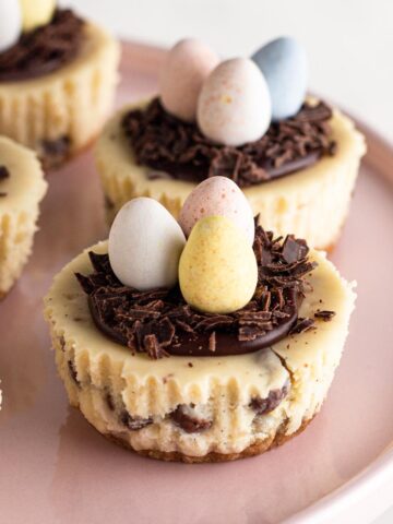 Mini cheesecakes topped with chocolate "nests" and Cadbury mini eggs on a pink cake stand.
