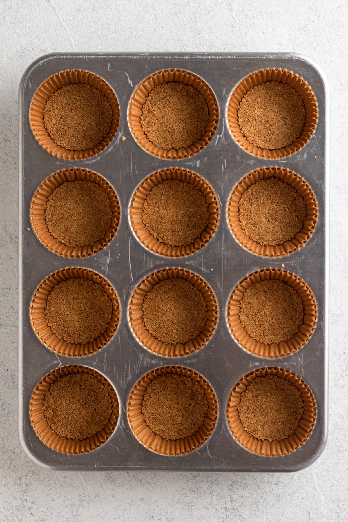 Baked graham cracker crusts in brown muffin liners in a muffin pan.