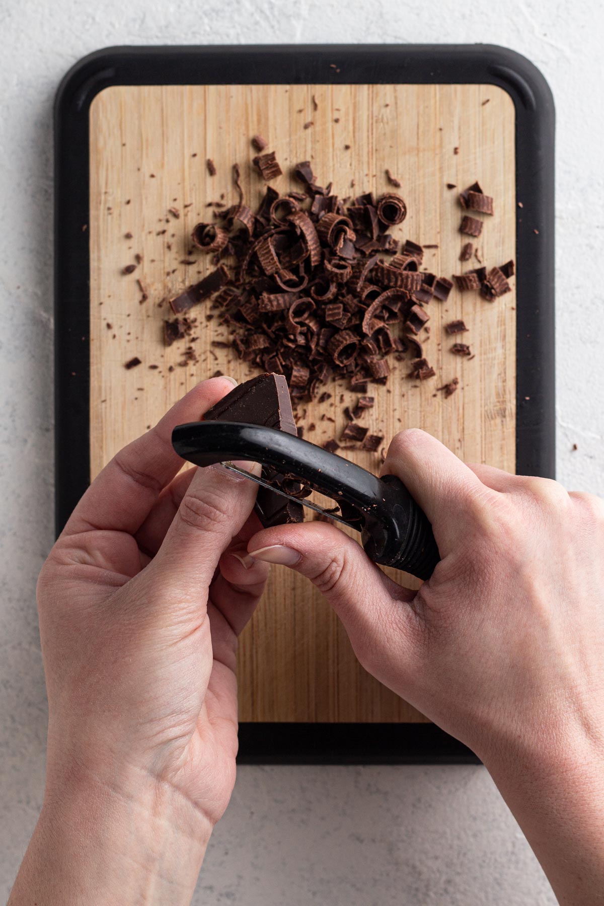 Hands holding a square of chocolate and a vegetable peeler, creating chocolate curls.