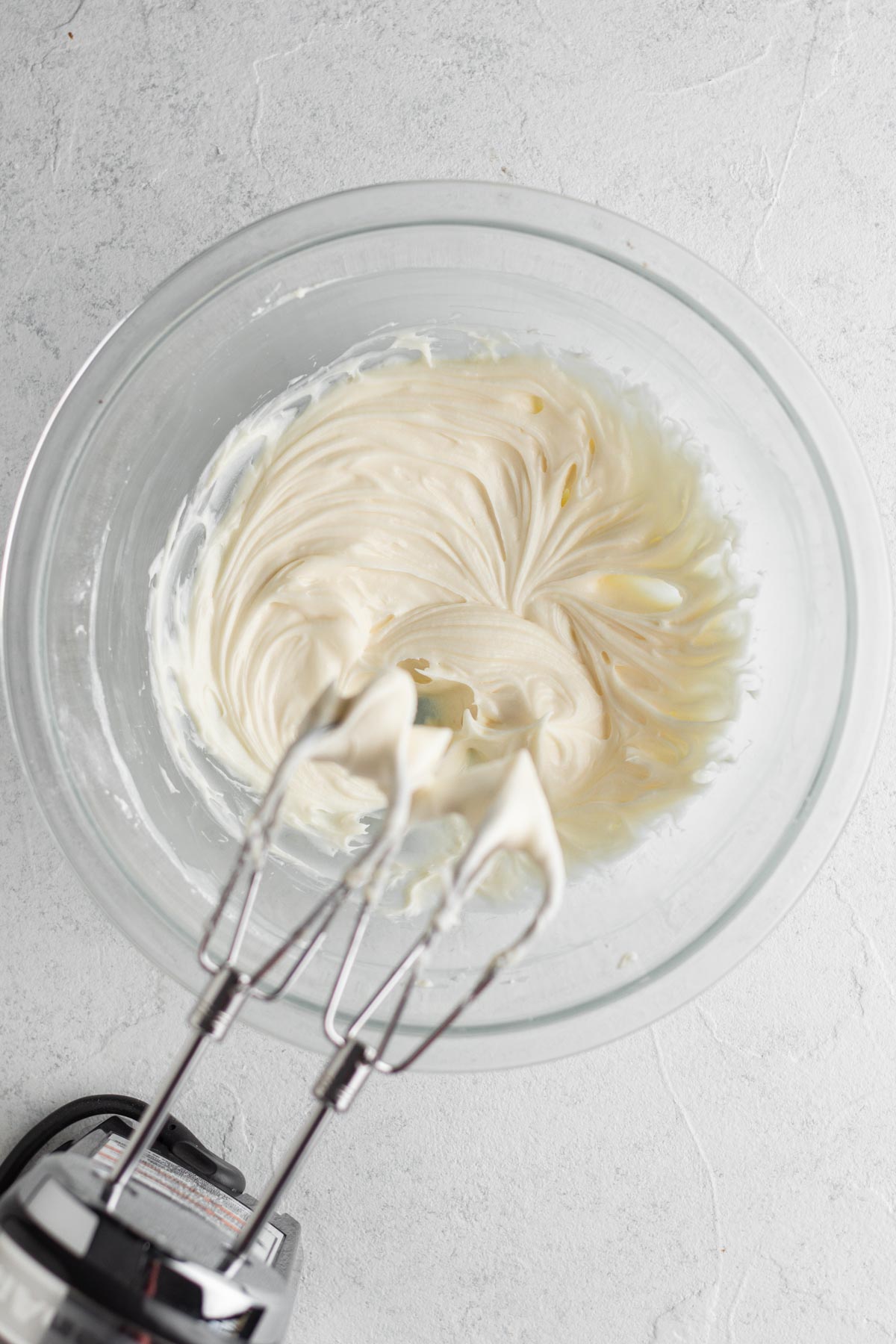Cream cheese frosting in a glass mixing bowl with an electric hand mixer.