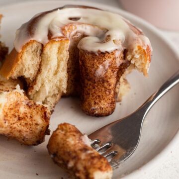 Angled view of an iced cinnamon roll cut up on a white plate with a fork.