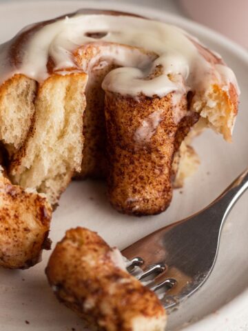 Angled view of an iced cinnamon roll cut up on a white plate with a fork.