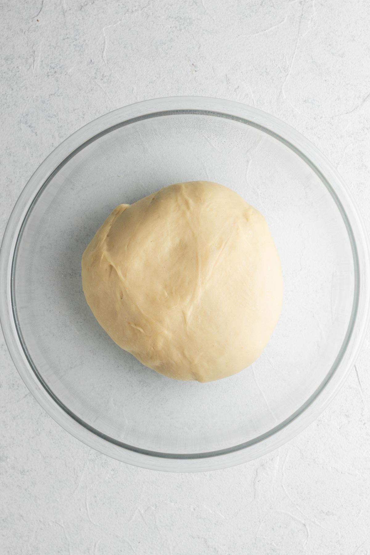 Ball of risen brioche dough in a glass mixing ball on a white surface.