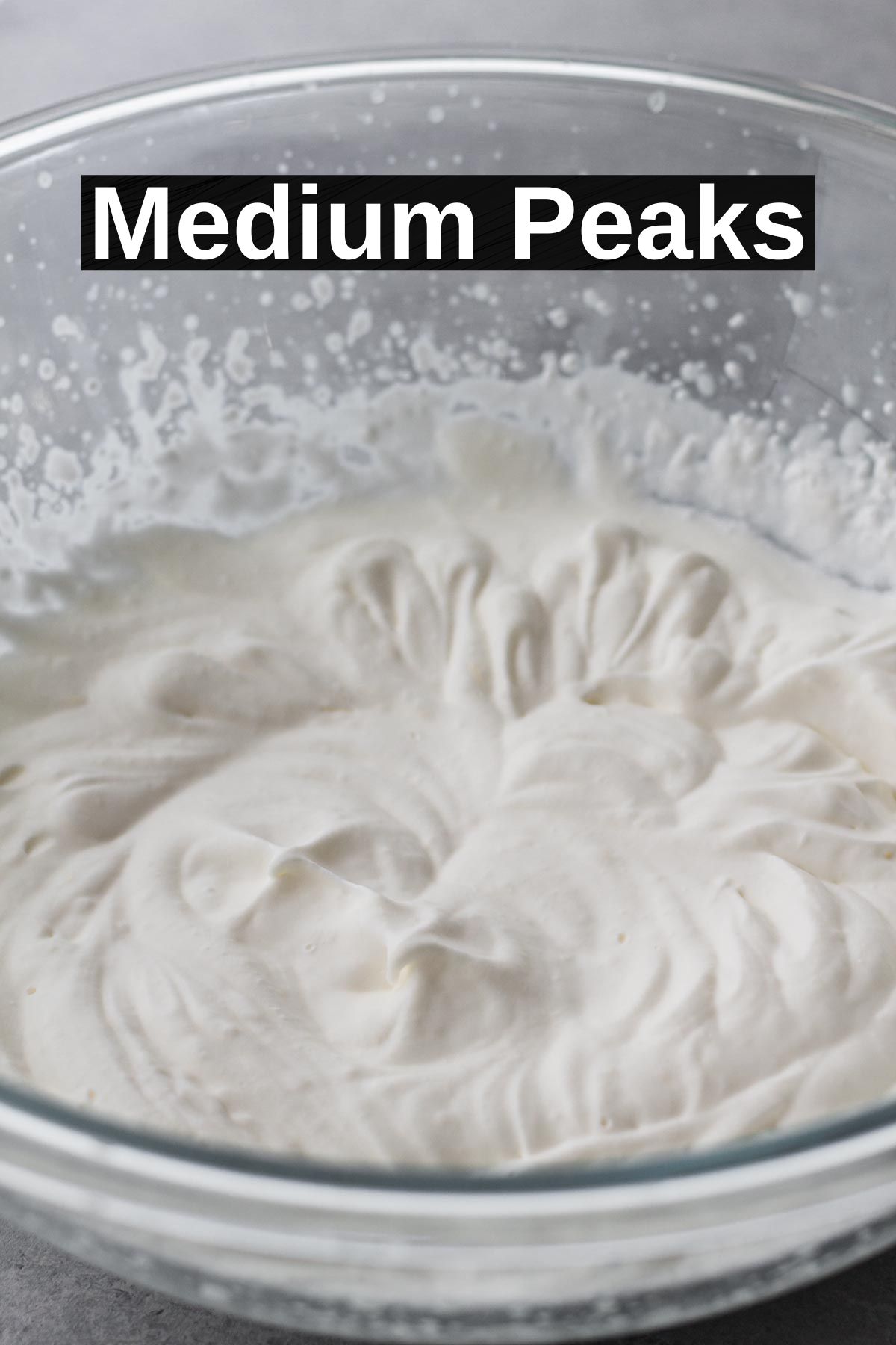 Glass bowl filled with medium peaks of whipped cream on a gray surface.