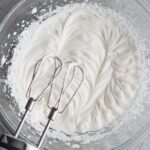 Overhead view of whipped cream in a glass bowl with an electric hand mixer.
