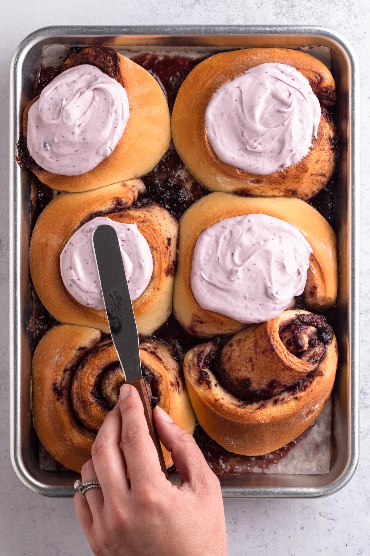 Hand using a spatula to spread icing over cinnamon rolls.