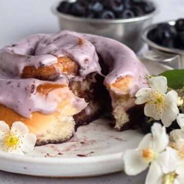 Close-up view of a cinnamon roll with purple icing split open on a white plate with a dish of blueberries and small white flowers.