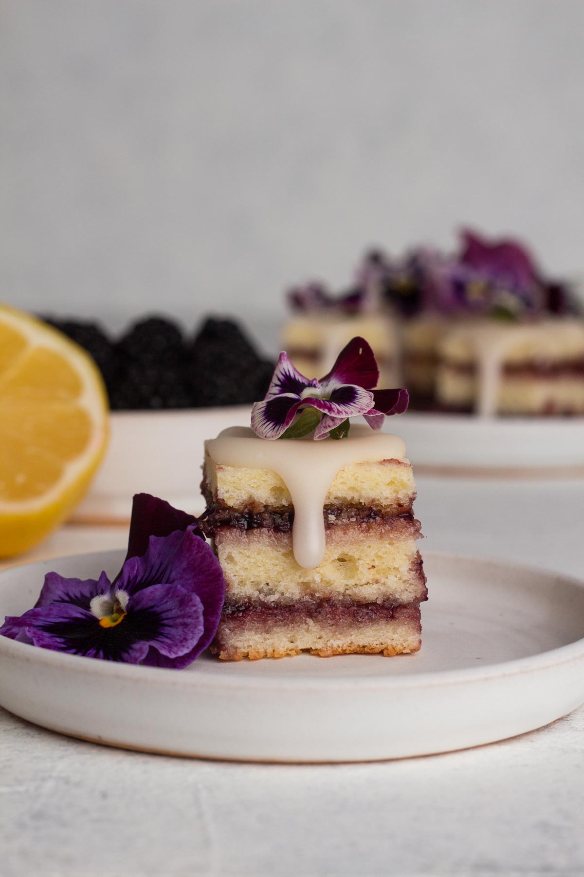 Side view of a petit four on a white plate topped with dripping glaze and purple pansy flowers.