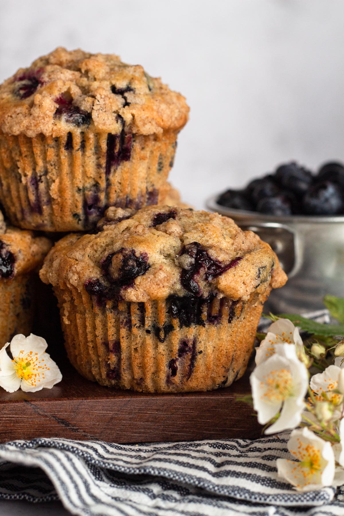 Blueberry muffins stacked on a wooden cutting board with white flowers and a cup of fresh blueberries.