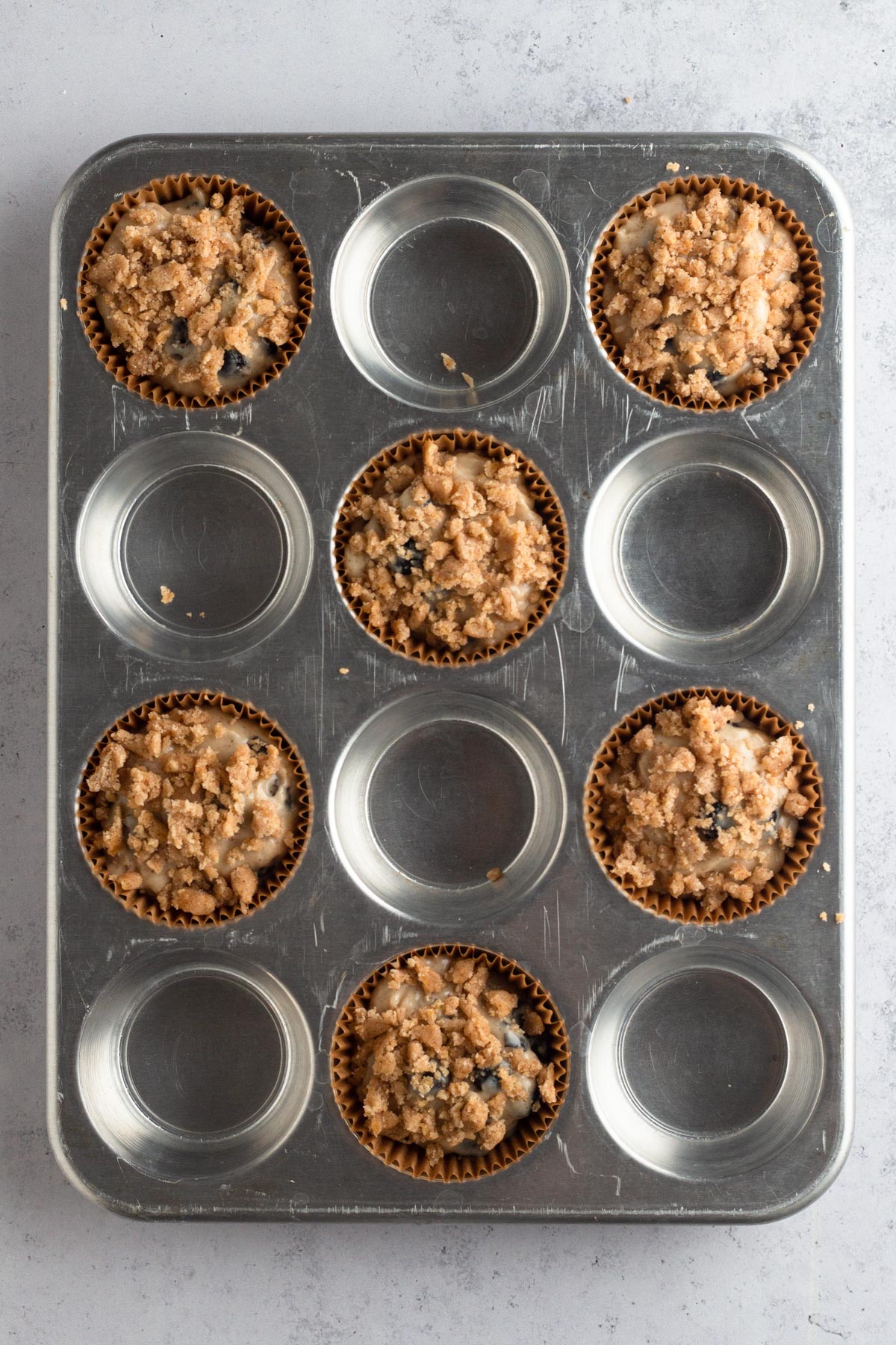 Muffin batter topped with streusel in brown paper liners in a metal muffin pan.