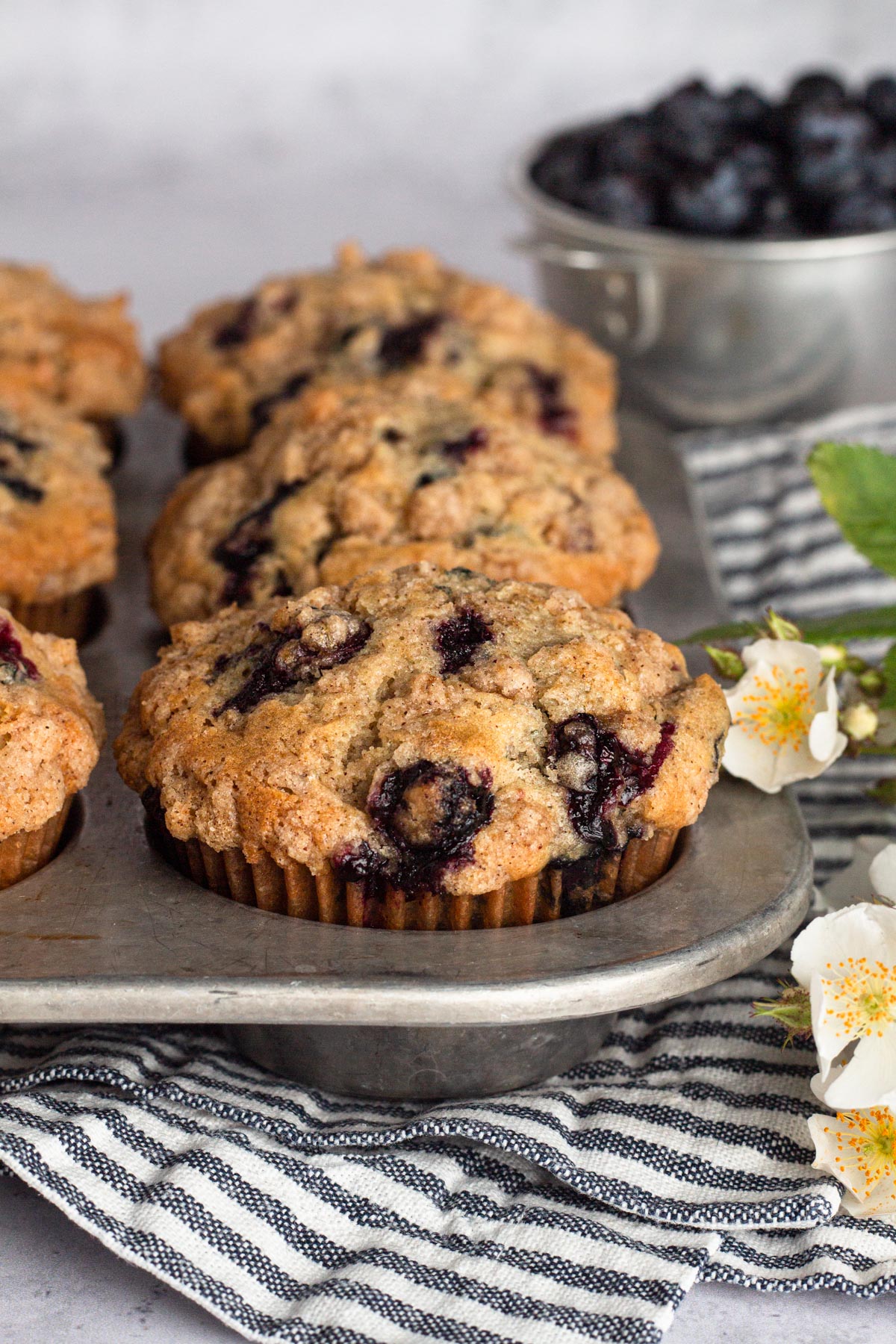 Angled view of blueberry muffins in a metal muffin tin with a striped blue napkin and white flowers.