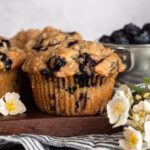 Blueberry muffins on a wooden cutting board with white flowers and fresh blueberries.