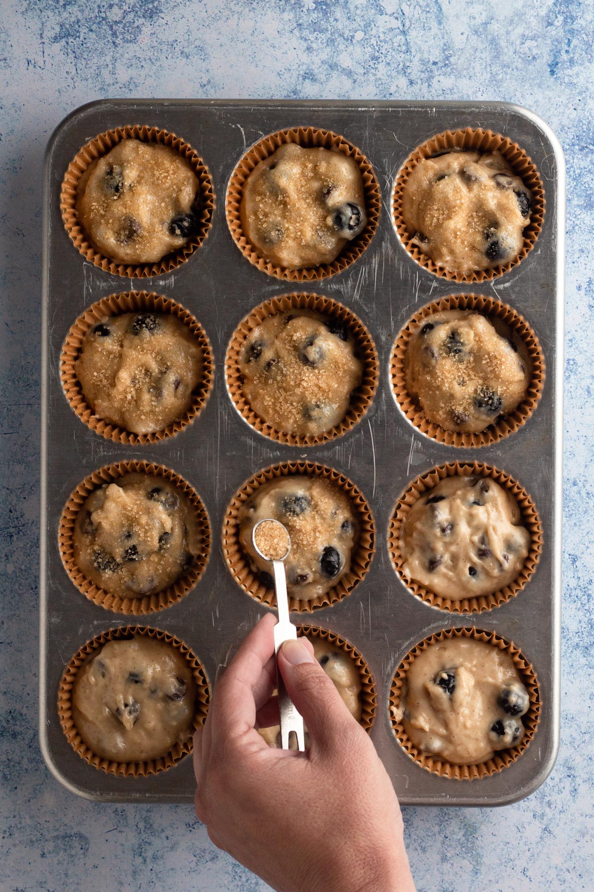 Hand holding a small measuring spoon and sprinkling sugar over muffin batter in pan.
