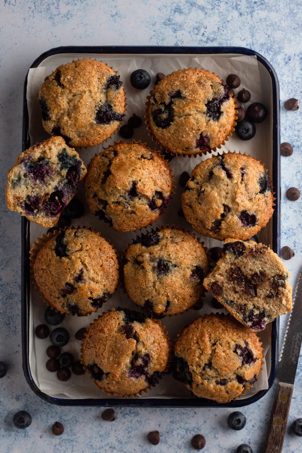 Overhead view of whole and sliced muffins on a rimmed tray surrounded by blueberries and chocolate chips.
