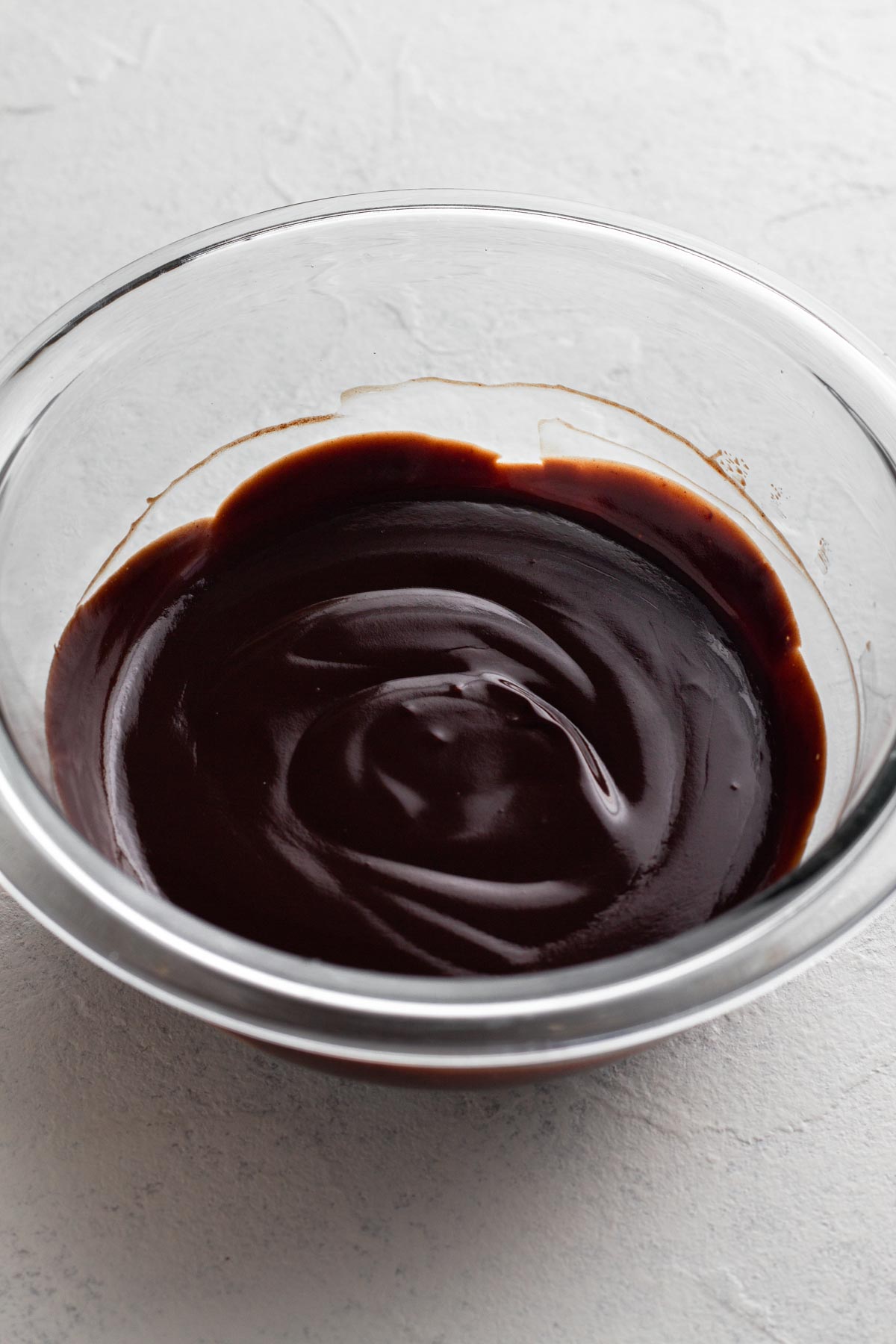 Angled view of chocolate ganache in a glass mixing bowl on a white surface.