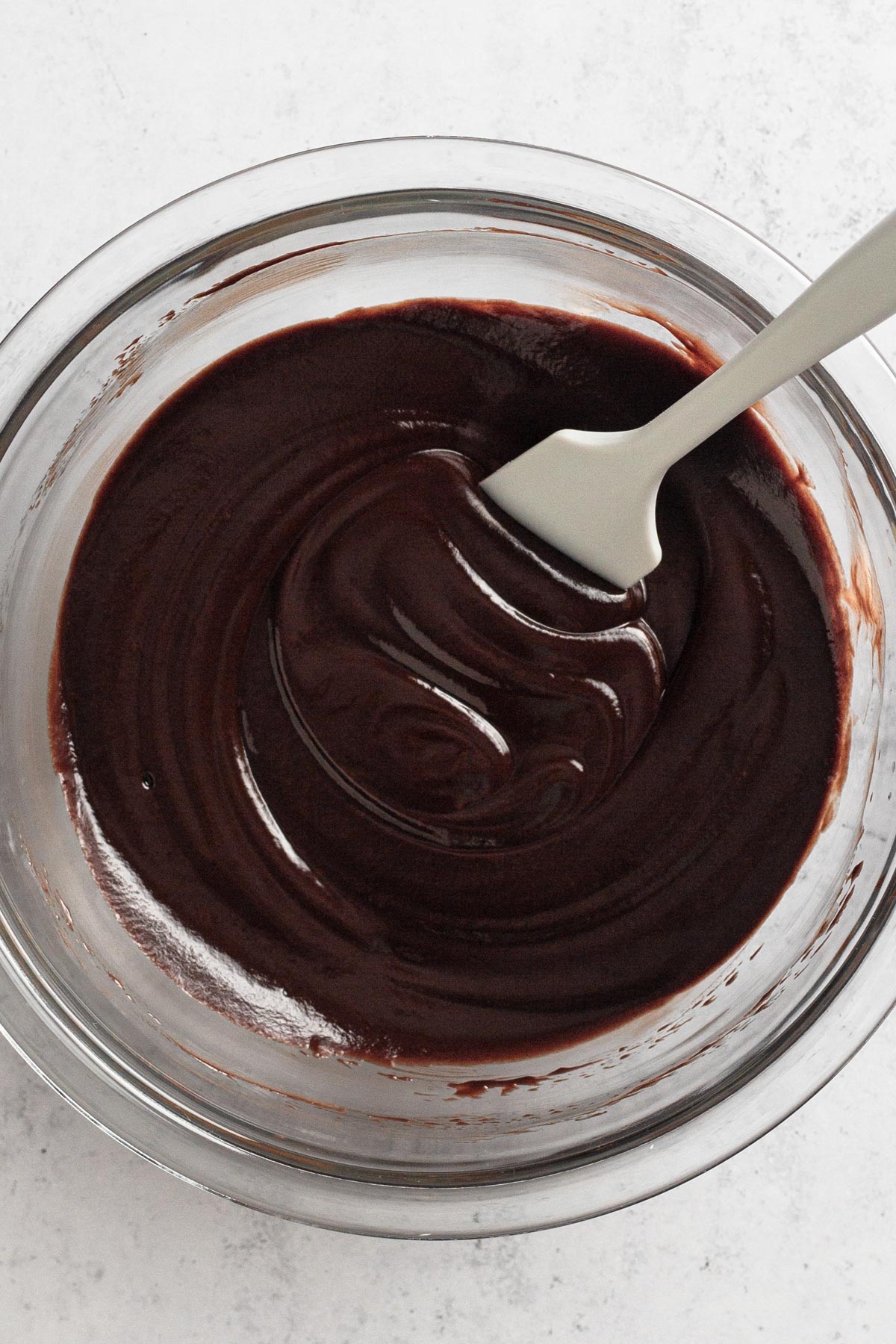 Overhead view of chocolate ganache in a glass mixing bowl with a white rubber spatula.