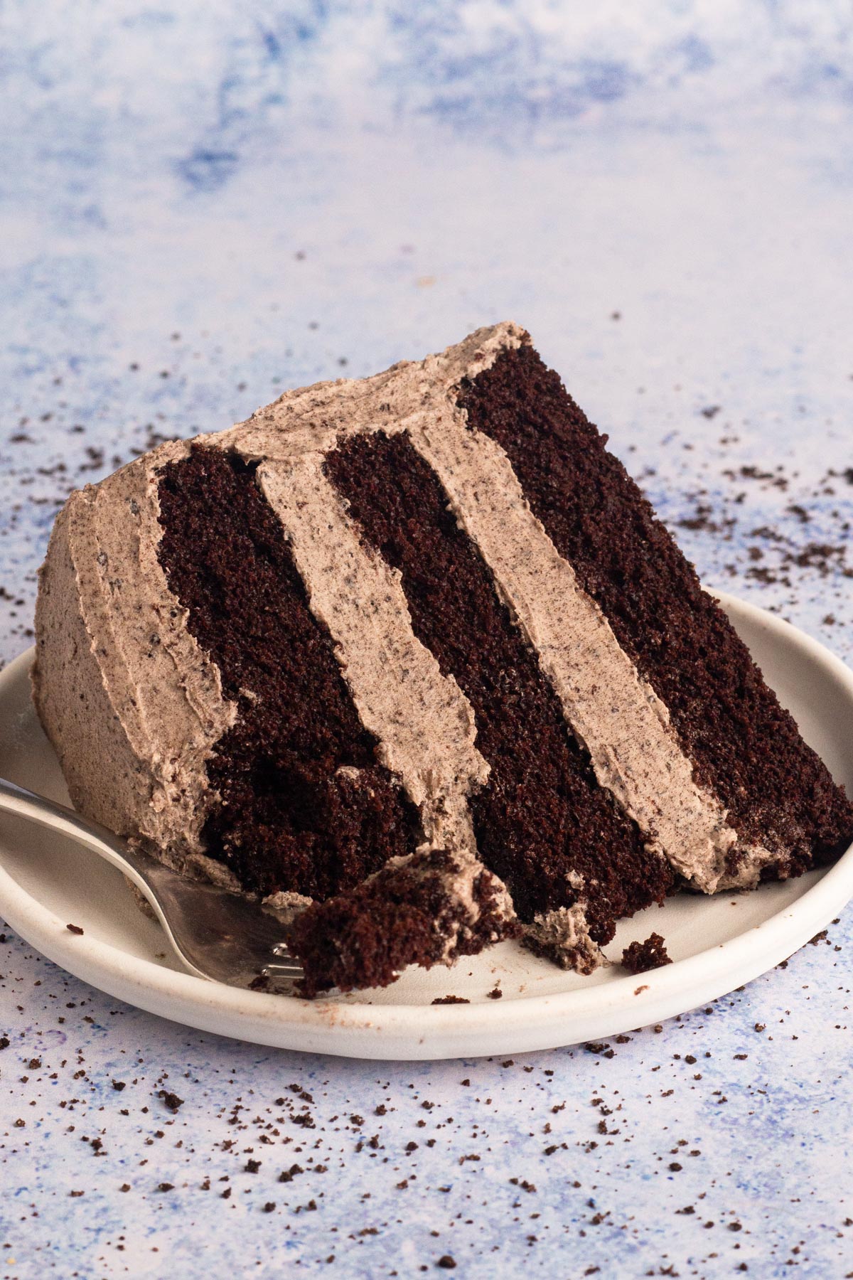A side view of a slice of chocolate layer cake with Oreo buttercream on a white plate. A fork is laying next to the cake on the plate holding a forkful of cake and frosting.