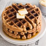 Angled view of stacked waffles with butter and maple syrup on a a plate with a newspaper.