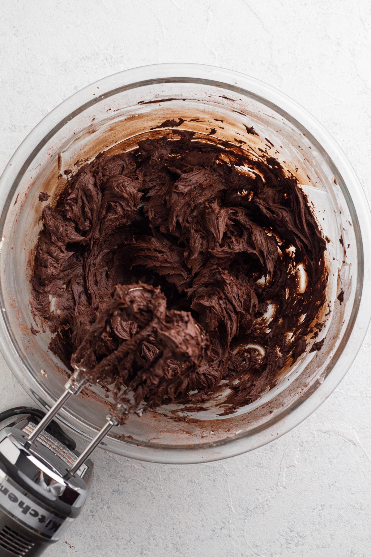 Chocolate buttercream frosting in a glass mixing bowl with an electric hand mixer.