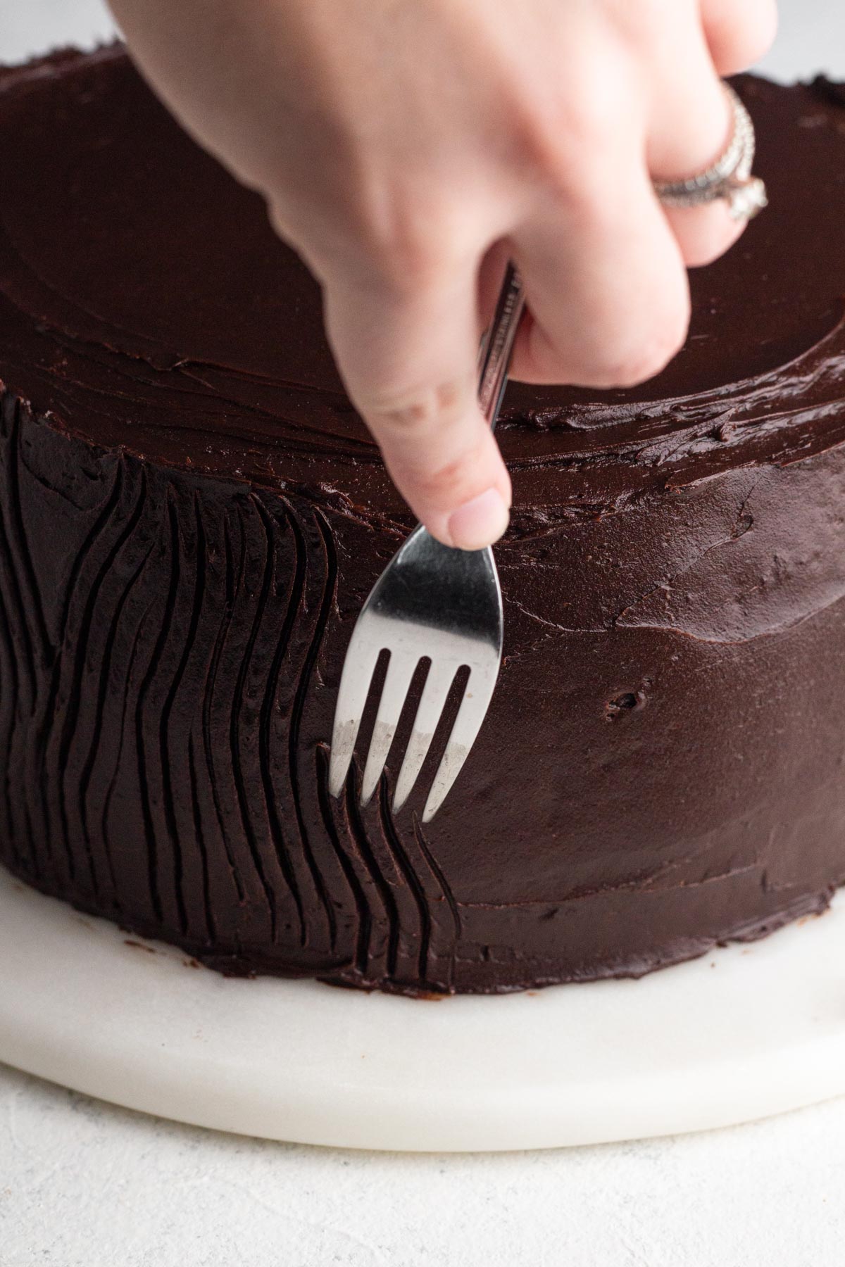 Hand holding a fork and dragging it through chocolate frosting to create a lined pattern in the frosting.