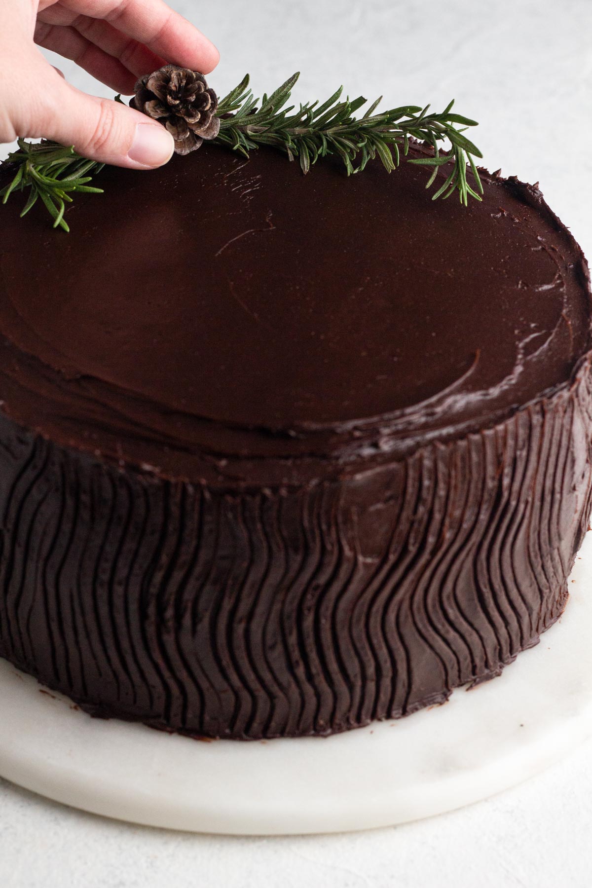 Hand placing a small pinecone onto the top of a chocolate-frosted cake garnished with sprigs of rosemary.