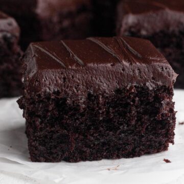 Close up view of a square of moist chocolate cake topped with chocolate frosting on a crumbled piece of parchment paper.