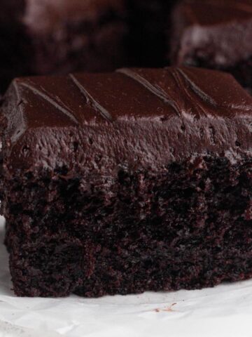 Close up view of a square of moist chocolate cake topped with chocolate frosting on a crumbled piece of parchment paper.