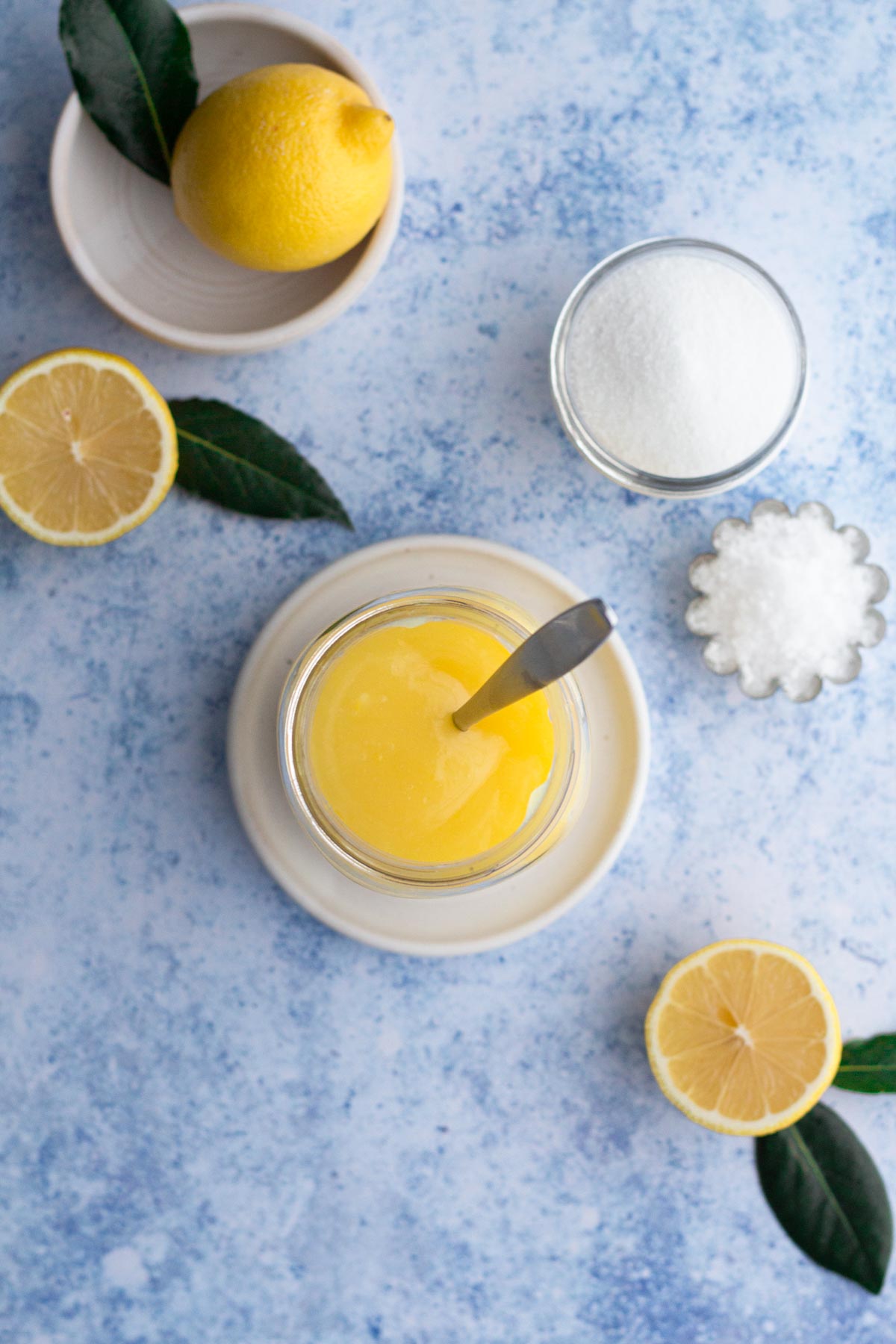 Overhead view of lemon curd in a glass jar surrounded by fresh lemons and sugar on a blue surface.