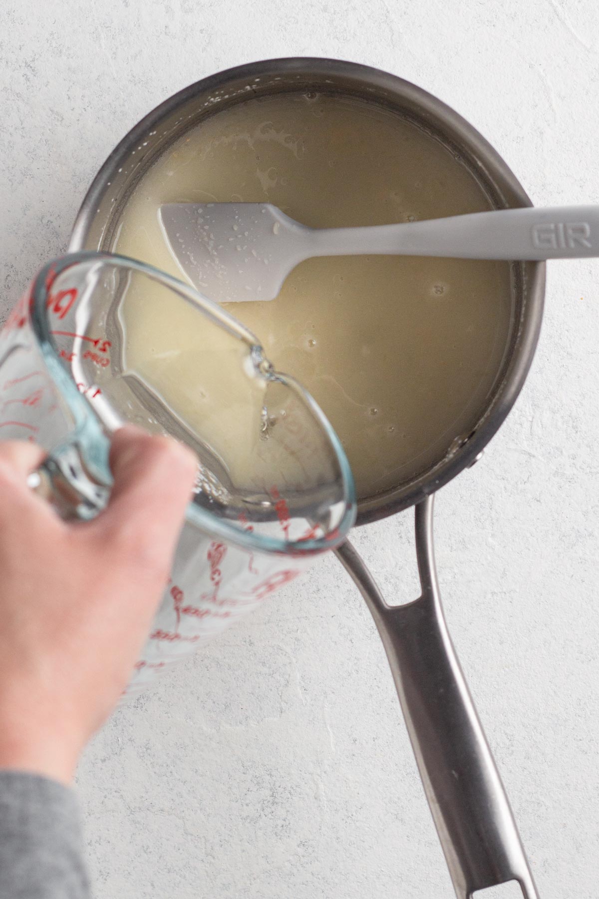 Hand pouring cold water and lemon juice into a saucepan with sugar and a white spatula.